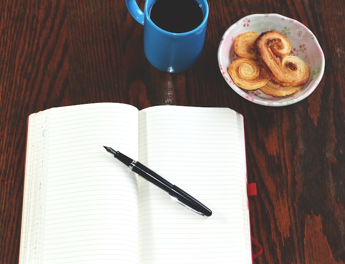 Offenes Notizbuch mit Stift neben Kaffee und Gebäck